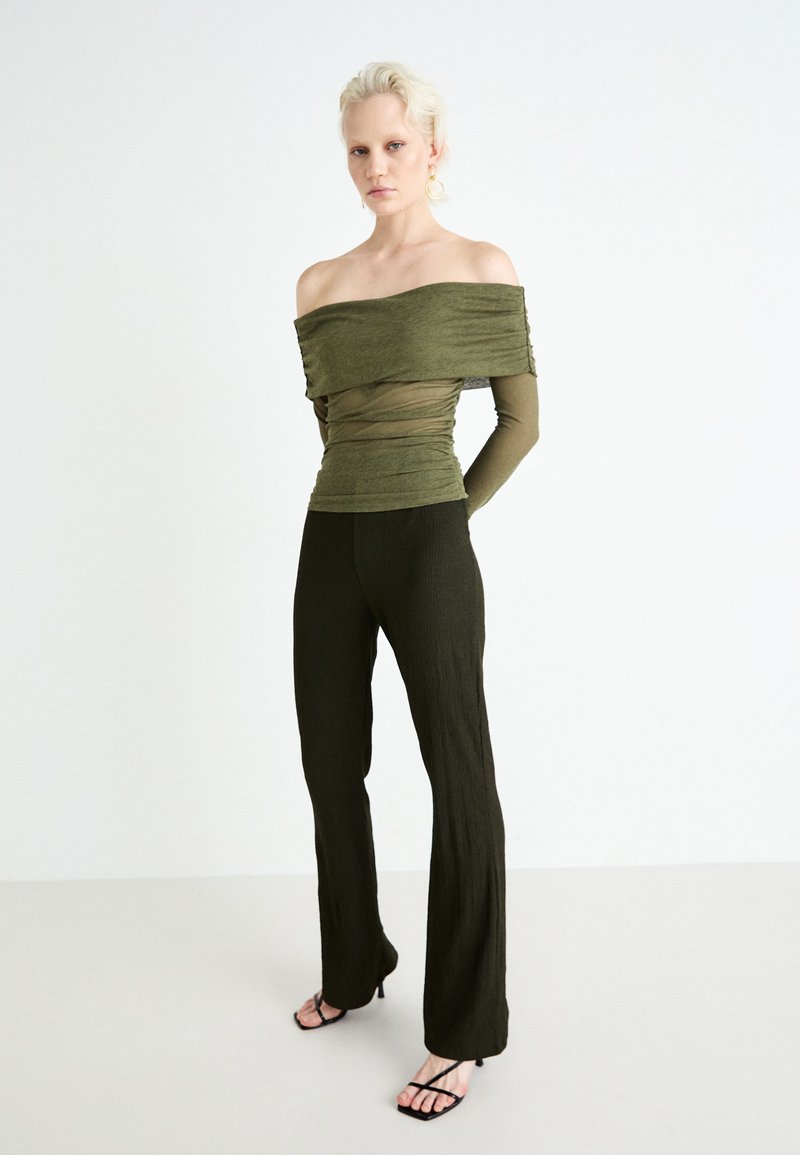 Olivengrünes Top mit schulterfreiem Schnitt, strukturiertem Stoff, schichtartigem Design und langen Ärmeln, kombiniert mit dunkelgrünen, gerippten Schlaghosen und schwarzen Sandalen.