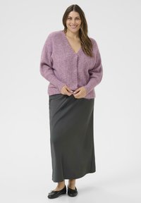 Glunderende vrouw in een roze V-hals trui, een lange donkere rok en zwarte ballerina's, staande tegen een eenvoudige witte achtergrond.