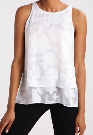 Haut blanc sans manches superposé avec motif jacquard floral porté par une personne debout, les mains détendues le long du corps.