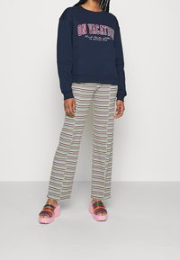 Person som bär en marinblå sweatshirt med texten "PÅ SEMESTER", randiga byxor med krusade kanter och rosa plattformssandaletter med flerfärgade remmar.