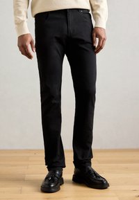 Μαύρο λεπτό παντελόνι από ελαφρύ ύφασμα, με κλασικό σχέδιο πέντε τσεπών και λείο υφή. Φορεμένο με μαύρα loafers.