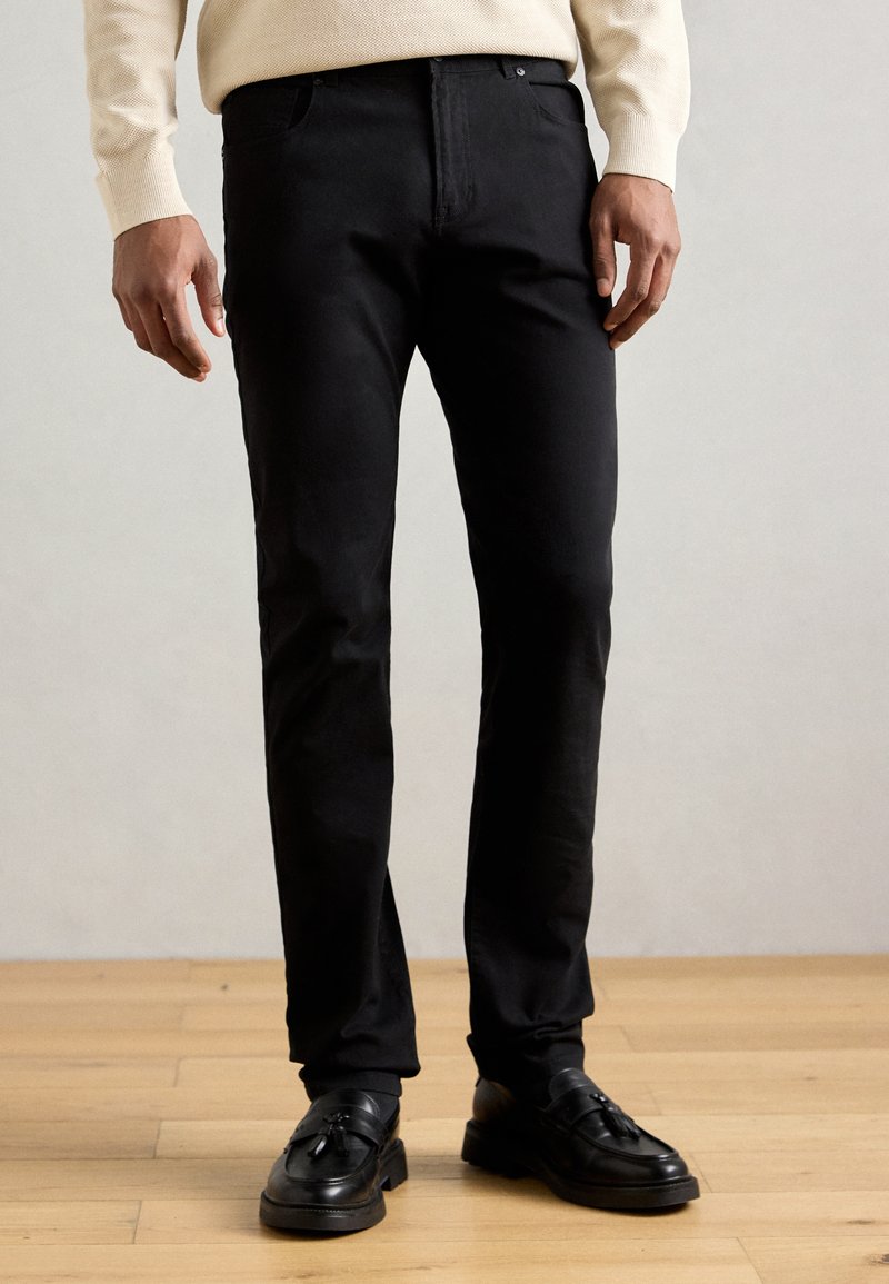 Μαύρο λεπτό παντελόνι από ελαφρύ ύφασμα, με κλασικό σχέδιο πέντε τσεπών και λείο υφή. Φορεμένο με μαύρα loafers.