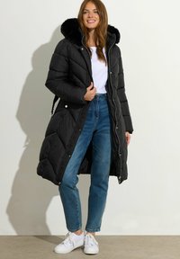 Sort pufferjakke med en faux fur hætte, lynlås, og sidelommer, brugt med lyseblå jeans og hvide sneakers.