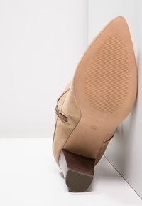 Botín de ante beige hasta el tobillo con punta afilada, tacón de madera estratificado y suela texturizada. Incluye una cremallera en el costado para facilitar su uso.