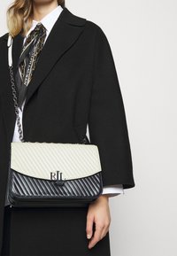 Quiltad handväska med en krämfärgad övre klaff och svart kropp, med en silverkedja som axelrem och präglad "RL"-logotyp på framsidan.