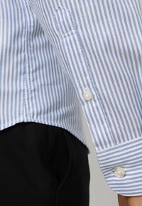 Ljusblå och vit randig skjorta med knappslutning och subtil struktur, ihopparad med svart tyg synligt nedanför, visad i en närbild.