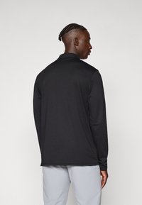 Μαύρο polo shirt με μακριά μανίκια, κατασκευασμένο από απαλό ύφασμα, που διαθέτει γιακά και ίσιο τελείωμα, με ελάχιστο σχεδιασμό και χωρίς ορατές λεπτομέρειες.