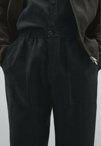 Pantalon noir avec une coupe décontractée, doté d'une taille élastique, de poches avant et d'un tissu texturé. Associé à une chemise à boutons foncée assortie.