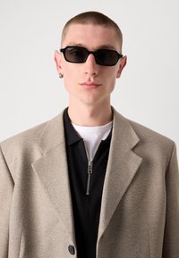 Blazer beige strutturato sopra una camicia nera con zip. Il modello indossa occhiali da sole ovali neri e un orecchino a cerchio in argento. Sfondo grigio semplice.