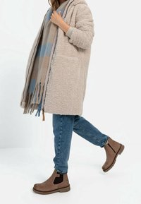 Bottines marron avec des panneaux élastiques sur les côtés, portées avec un jean bleu et un manteau en fausse fourrure beige, drapées avec une écharpe en plaid.