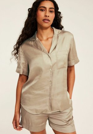 Vrouw met lang krullend haar, gekleed in een lichtbeige kort mouwen overhemd met knopen en bijpassende korte broek tegen een effen achtergrond.
