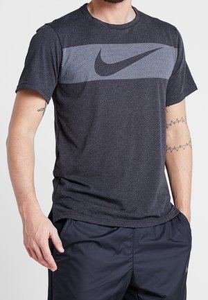 Mann trägt dunkelgraues Nike-T-Shirt mit Logo über der Brust und schwarze Sportshorts, steht mit den Händen nahe an den Taschen.