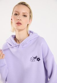 Lyselilla hettegenser med lommer foran og snor. Har svarte trykte design av kirsebær, en sommerfugl og en blomst. Myk tekstur.