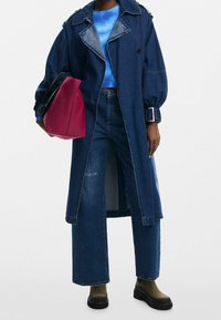 Personne portant un jean large en denim et un long manteau en denim, tenant un grand sac à main rouge, et portant des chaussures plateformes noires.
