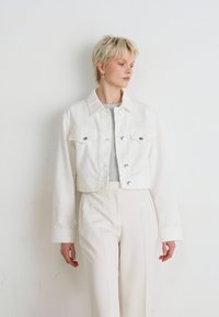 Cropped white denim jacket with silver buttons and four pockets, worn over a light gray top, paired with high-waisted white trousers.