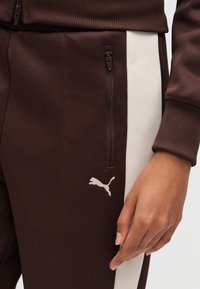 Καφέ αθλητικό παντελόνι με υφαντό ύφασμα, με λευκή πλευρική ρίγα, τσεπάκι με φερμουάρ και μικρό λευκό λογότυπο Puma στο πόδι.
