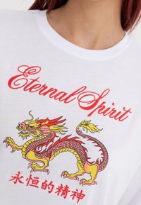 Hvid bomulds t-shirt med røde broderede bogstaver "Eternal Spirit" og et farverigt dragegrafik i gule, røde og pink nuancer.