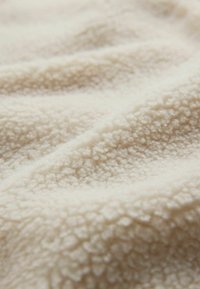 Mjukt, texturerat fleece i en ljus gräddfärg, med vågor och fördjupningar över ytan som framhäver dess plyschiga och mysiga känsla.