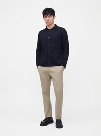 Jeune homme debout, les mains dans les poches, portant une chemise sombre boutonnée, un pantalon beige et des chaussures noires sur fond blanc.