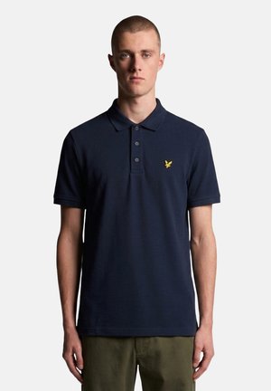 Jonge man met kort haar, die een donkerblauw poloshirt met een geel vogel-logo en olijfgroene broek draagt, staat voor een witte achtergrond.