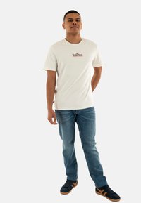 Camisa de algodón blanca de manga corta con un logo marrón de Timberland, combinada con jeans de denim azul y zapatillas negras.