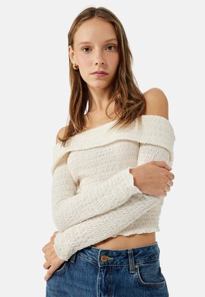 Pull court blanc à épaules dénudées avec un motif en vagues texturé, associé à un jean en denim taille haute avec fermeture à bouton.
