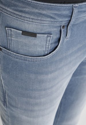 Plave traperice s uskim krojem, svijetlim pranjem i stražnjim džepom s crnom etiketom. Tkanina prikazuje suptilnu teksturu i detalje šivanja.