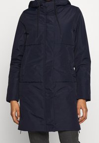 Mörkblå vadderad jacka med huva, långa ärmar, frontdragkedja, två stora fickor och snörningar vid huvan och nederkanten. Texturerad tyg.