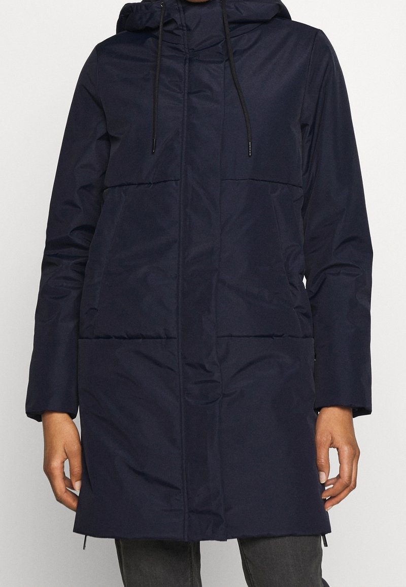 Mörkblå vadderad jacka med huva, långa ärmar, frontdragkedja, två stora fickor och snörningar vid huvan och nederkanten. Texturerad tyg.