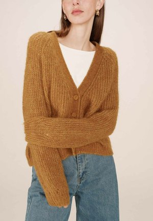 Cardigan tricoté de couleur moutarde chaude, avec un col en V, deux boutons, et un tricot doux et texturé. Assorti avec un jean bleu clair.