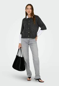 Jeune femme portant un cardigan gris foncé à boutons sur une chemise blanche, un jean gris clair, des sandales à talons noires, tenant un grand sac fourre-tout noir.
