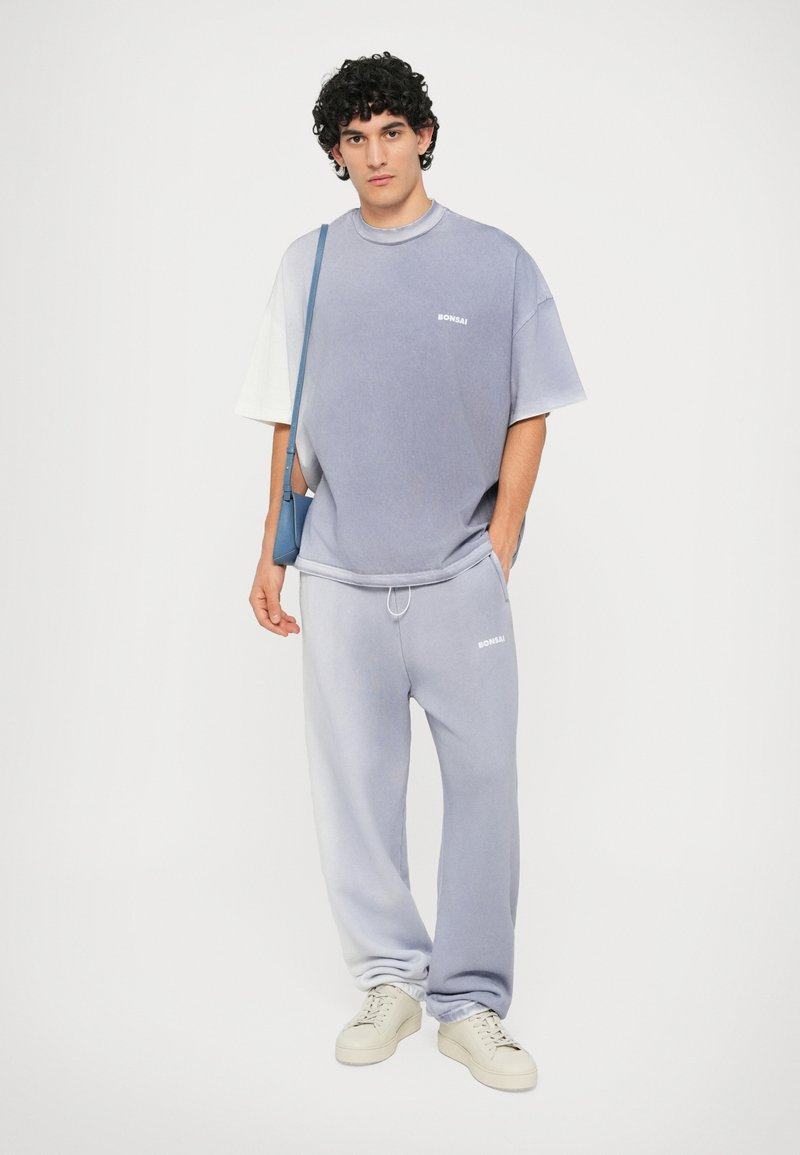 Tricou oversized lavandă cu accente albe și logo, completat cu pantaloni de trening tapered asortați. Tenisi albi finalizează ținuta.