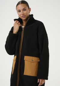 Zwarte fleece-jas met hoge kraag, voorzien van lichtbruine stoffen zakken en een ritssluiting aan de voorkant. De textuur is zacht en pluizig.