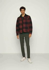 Red and black checkered jacket with two chest pockets, worn over a dark green shirt, paired with gray pants and white sneakers.