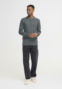 Grijze gebreide trui met een kraag en drie knopen, gecombineerd met donkere jeans en lichte sneakers, staand tegen een effen achtergrond.