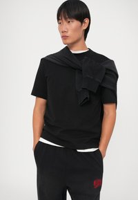 Svart bomulls T-skjorte med korte ermer, kombinert med svarte joggebukser som har en rød logo. Genser over skuldrene.