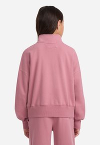 Rosa genser med høy krage, senkede skuldre og ribbestrikkede mansjetter og kant. Har et merke ved nakken og myk, teksturert stoff.