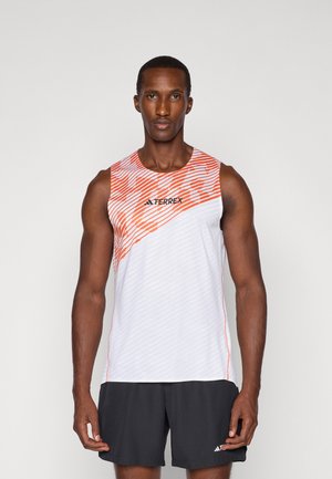 Aktiivne mees, kes kannab valget ja oranži ilma varrukateta TERREX särki ning musti Adidas lühikesi pükse, seisab sirgelt, nägu otse kaamera poole, lihtsa tausta ees.