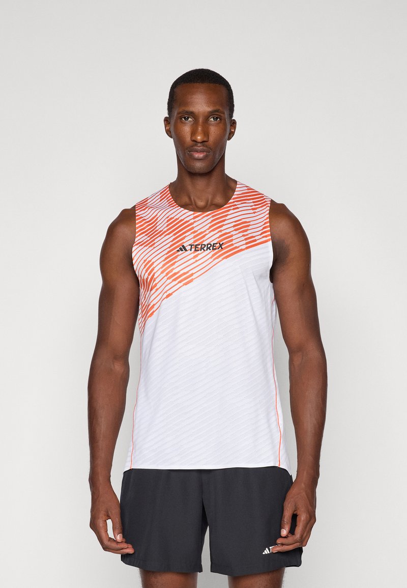 Mężczyzna sportowy w białej i pomarańczowej koszulce bez rękawów TERREX oraz czarnych szortach Adidas stoi przodem do aparatu na jednolitym tle.