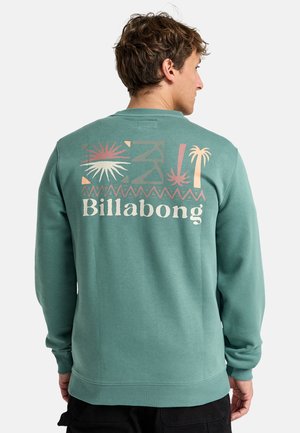 Grüne Sweatshirt mit langen Ärmeln, das auf der Rückseite einen großen Graf print mit dem Markennamen "Billabong" und tropischen Motiven aufweist.