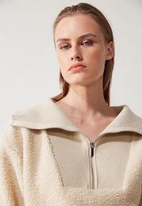 Giacca beige con zip, caratterizzata da una texture morbida e pelosa, con colletto a costine e dettaglio della zip. Il design complessivo è minimalista e contemporaneo.