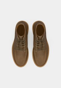 Un paio di stivali in suede marrone con lacci, punta tonda e cuciture visibili, visti dall'alto su uno sfondo bianco.