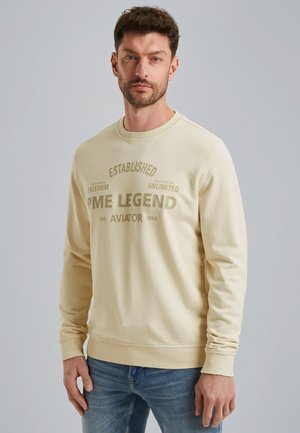 Sweat-shirt crème à manches longues, encolure ronde et impression en relief. Associé à un jean bleu. Design simple et décontracté.