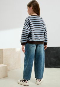 Barn står med ryggen vänd, klädd i en blå och brun randig tröja, lösa blå jeans och vita hög-top skor med svarta och röda mönster.