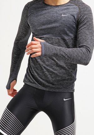 Person trägt schwarze und graue Nike-Sportbekleidung mit Daumenlöchern im Langarmshirt und posiert in Bewegung vor weißem Hintergrund.