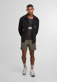 Młody mężczyzna stojący w casualowym ubraniu, czarna koszula, ciemny podkoszulek, oliwkowe szorty, beżowe skarpety i białe sneakersy na jednolitym tle.