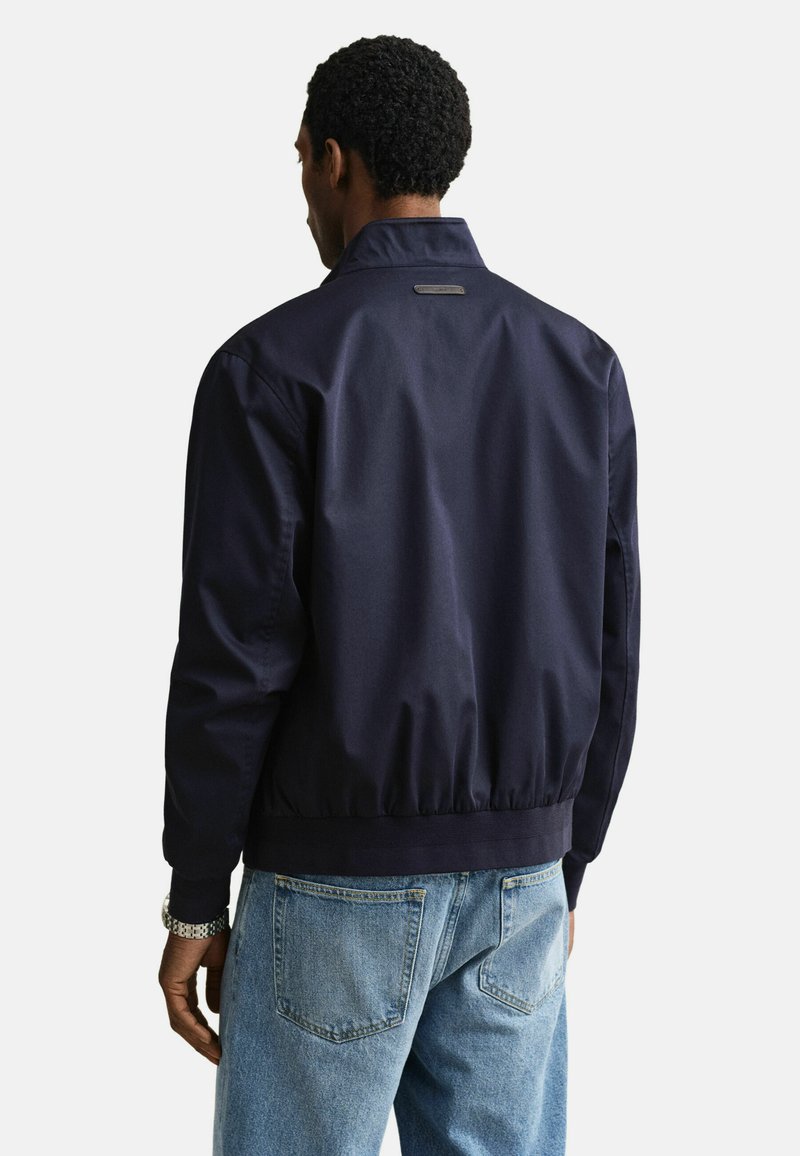 Homme portant une veste bleu marine et un jean bleu clair, vu de dos sur un fond uni.