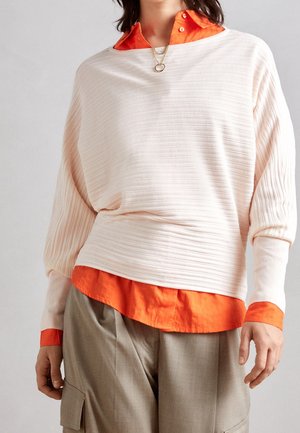 Personne portant un pull côtelé écru superposé à une chemise orange boutonnée avec un pantalon cargo beige.