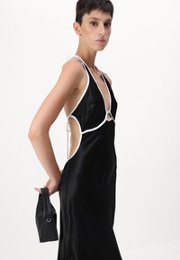 Black satin dress featuring a deep V-neck, white trim, open sides, and a back tie detail. Model holds a small black handbag.
