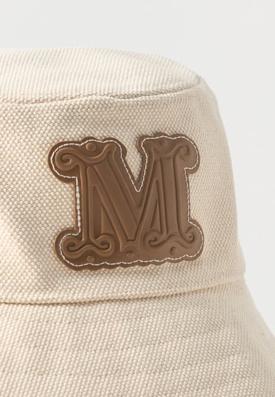 Μπεζ υφασμάτινο καπέλο τύπου κάδος με κεντημένο καφέ δερμάτινο ταμπελάκι που φέρει μπροστά ένα μεγάλο, περίτεχνο γράμμα «Μ».